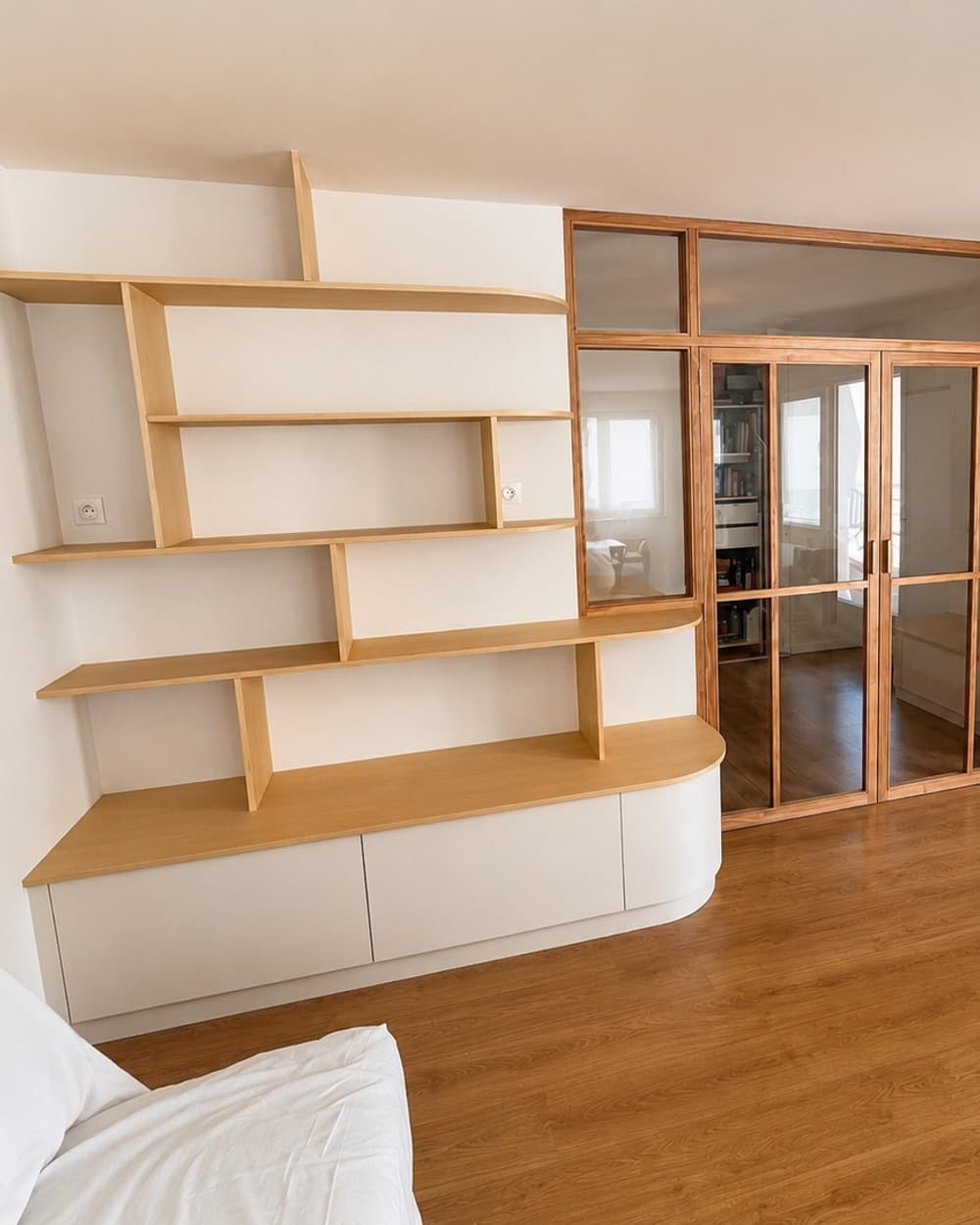 Côté salon, la bibliothèque aux étagères décalées sur un meuble bas arrondi, à côté de la verrière en frêne